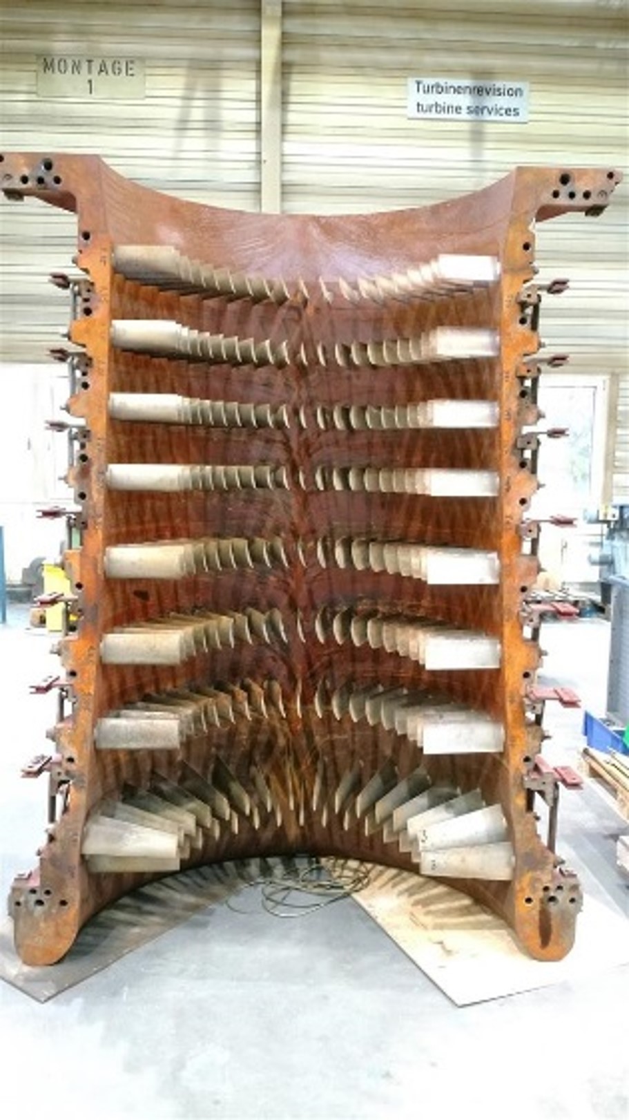  Large, cylindrical industrial component with multiple layers of metal compounds arranged in a regular pattern around the structure. The component is red and stands in a factory environment surrounded by other machinery and equipment. The background shows shelves and tools that indicate industrial use.