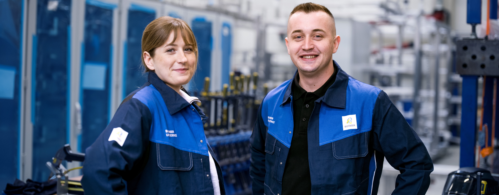 This image shows two employees in Bilfinger uniforms standing in an industrial setting, underscoring teamwork and expertise in engineering and manufacturing.