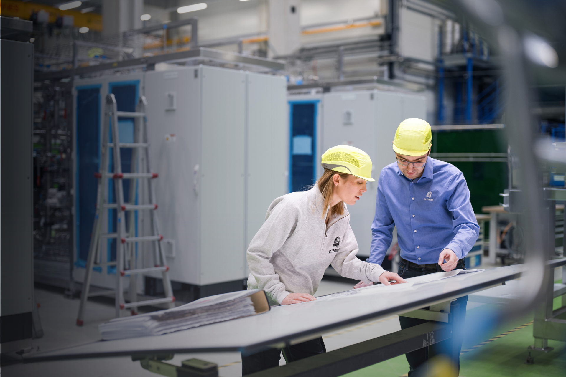 Das Bild zeigt zwei Personen mit gelben Schutzhelmen und Kleidung mit Bilfinger-Logo, die sich in einer industriellen Umgebung über Dokumente auf einem Tisch unterhalten oder diese überprüfen.