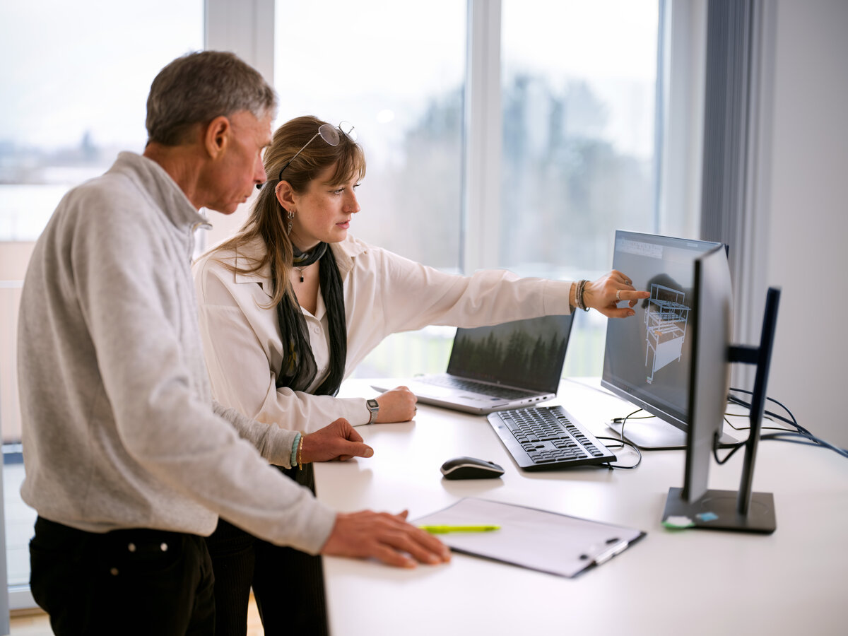 This image is a photograph depicting a collaborative work environment where two individuals are engaged in discussing a design or technical drawing displayed on a computer screen. The setting emphasizes teamwork and the use of technology in professional settings.