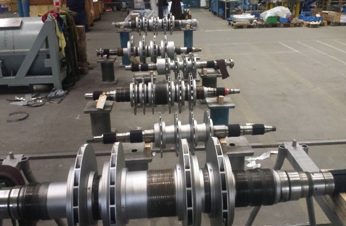 Industrial workshop with large metallic machine parts, shafts, and rotors on workbenches, surrounded by machines and tools.