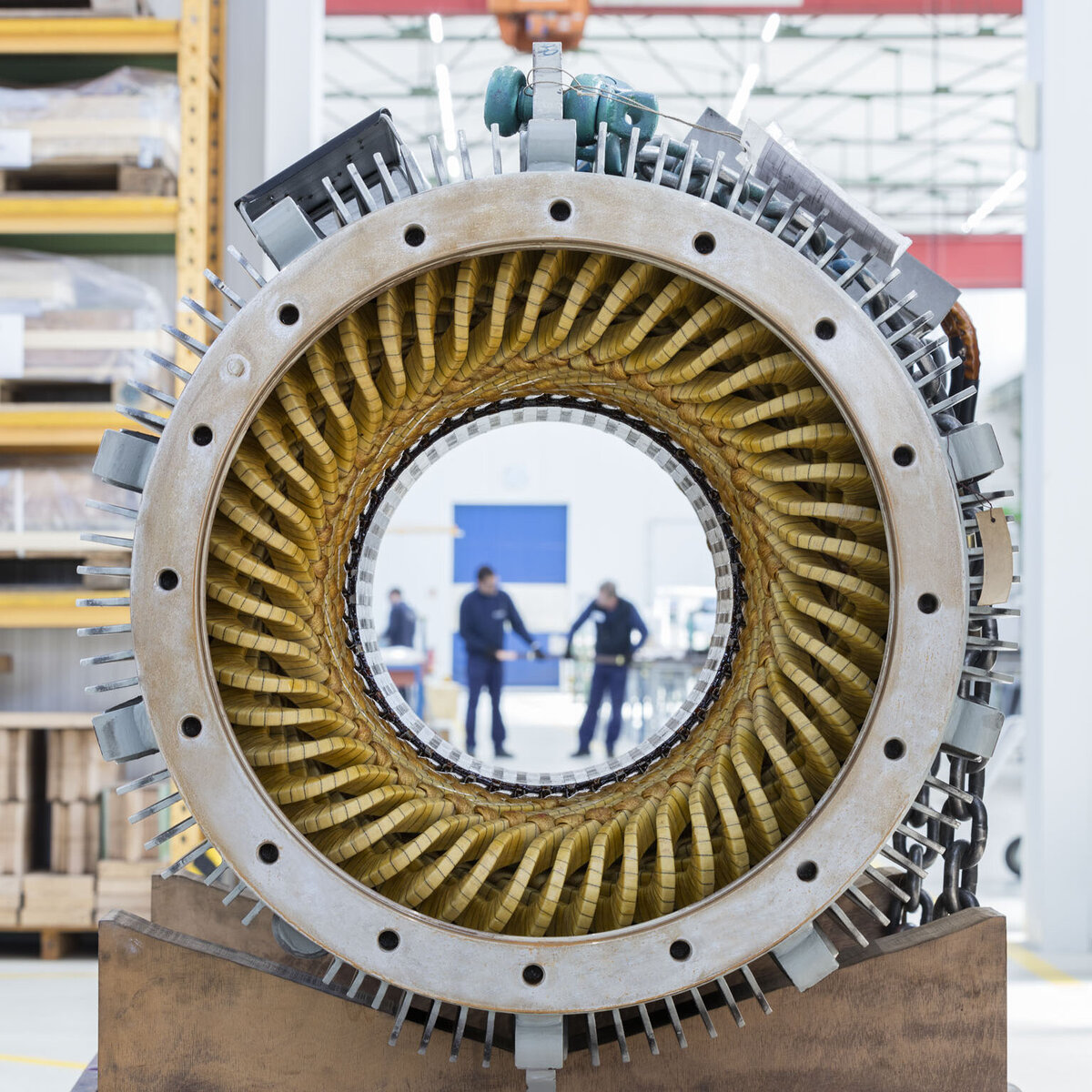Front view of a large electric motor stator with yellow windings arranged in a circular pattern. Through the center of the stator, two workers can be seen talking to each other in a bright factory environment. In the background, industrial shelving and a crane labeled “ABUS 5t” are visible, emphasizing the size and functionality of the production facility.