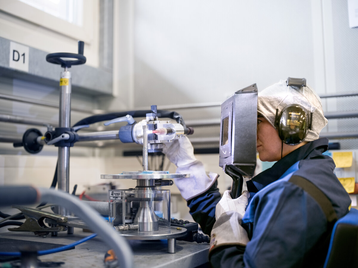 This image is a photograph of a female welder performing a technical task with specialized equipment in an industrial setting, highlighting the skilled nature of her work. The focus on machinery and protective gear underscores the precision and safety measures essential in such environments.