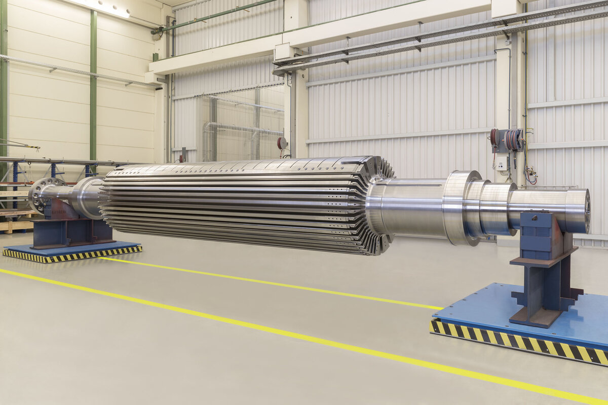 Large industrial metal roller in a modern factory hall. The roller is mounted on blue stands and surrounded by yellow safety lines. Industrial equipment and cables are visible in the background.