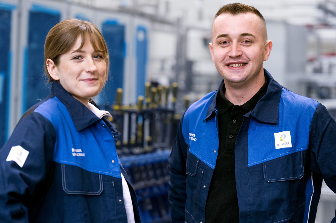 This image shows two employees in Bilfinger uniforms standing in an industrial setting, underscoring teamwork and expertise in engineering and manufacturing.