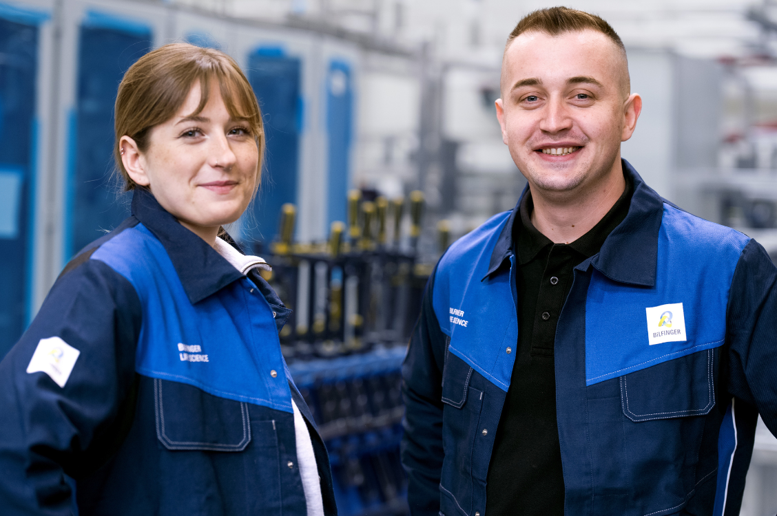 This image shows two employees in Bilfinger uniforms standing in an industrial setting, underscoring teamwork and expertise in engineering and manufacturing.