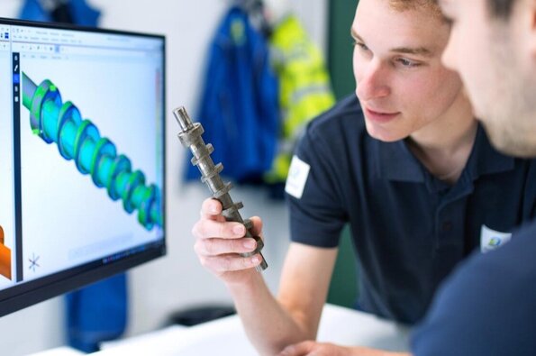 This image depicts Bilfinger employees engaged in engineering work, showcasing a computer screen with 3D models of a mechanical component and an employee holding a physical version of the component, emphasizing the integration of digital design and physical engineering.