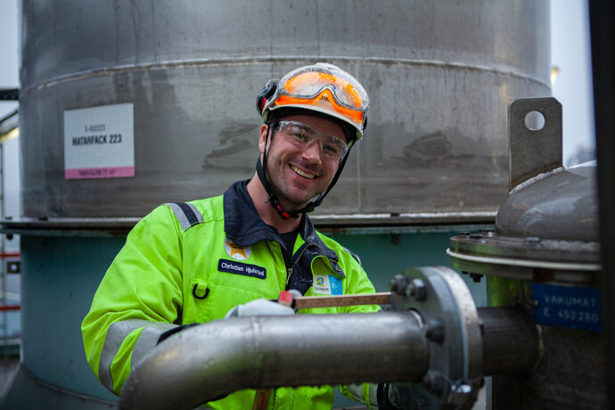 Male colleague standing in front of a pipe at a factory smiling at the camera. He is wearing a yellow overall, safety goggles and a white safety helmet.