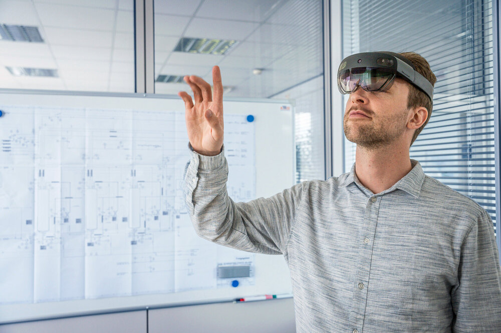 Reference_augmented Reality Person wearing a virtual reality headset interacting with virtual objects in a modern office with technical diagrams in the background.