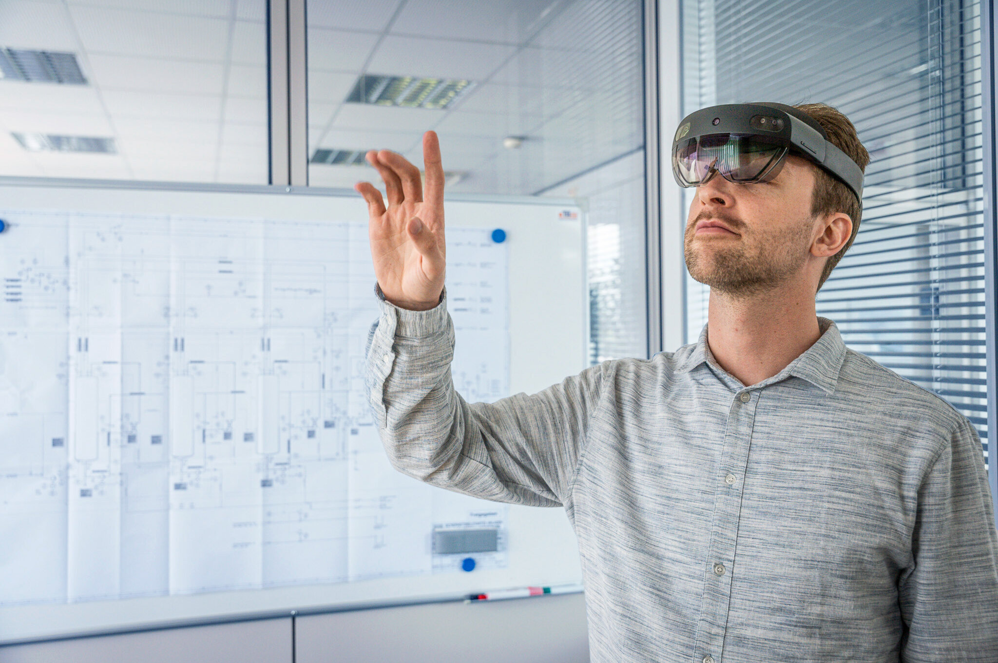 Person wearing a virtual reality headset interacting with virtual objects in a modern office with technical diagrams in the background.