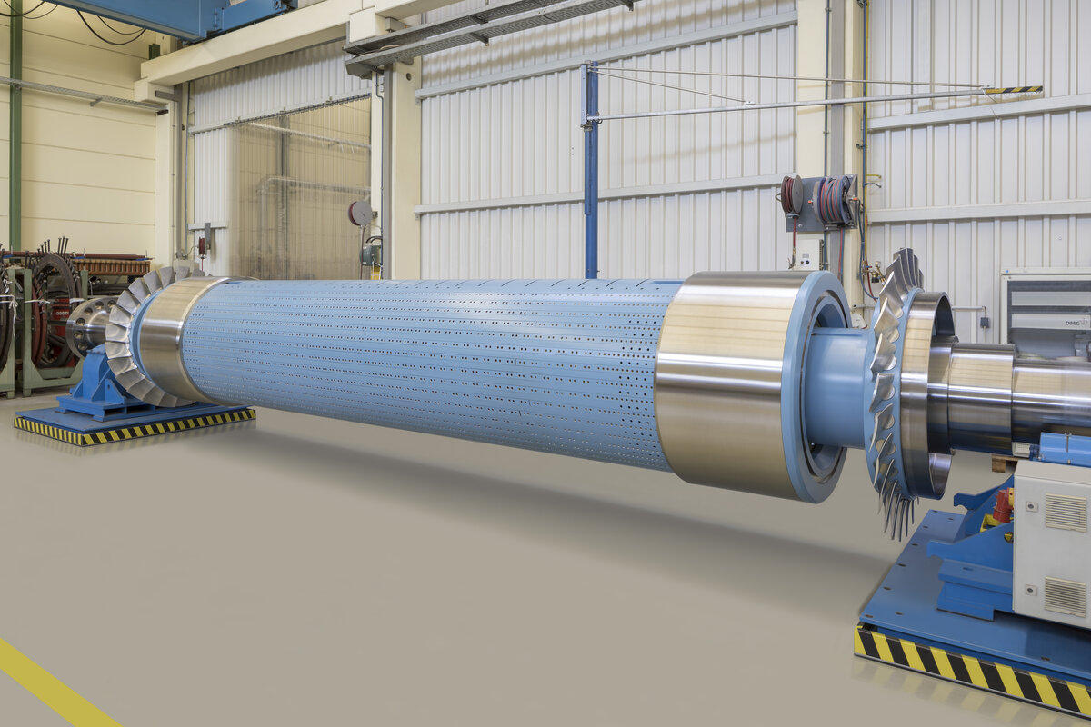 Large industrial rotor in a workshop or manufacturing facility. The rotor is cylindrical with a blue perforated surface and metallic ends. It is mounted on supports at both ends, with one end featuring blades or fins. The background includes various industrial equipment and machinery, as well as large doors and cables.