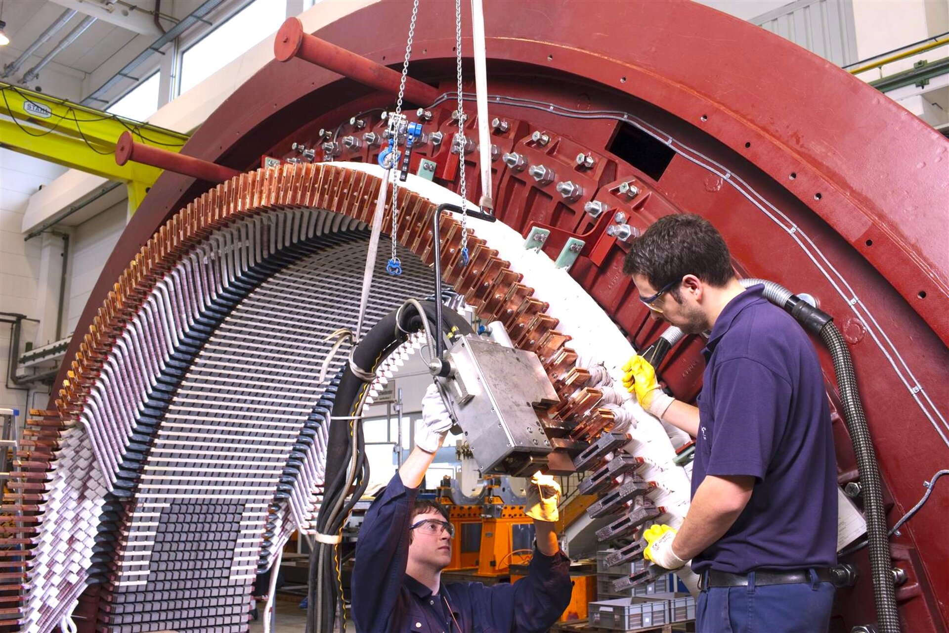 Two individuals working on an 18-pole motor/generator by AEG with a capacity of 35 MVA. The machine has a large circular structure with numerous copper coils and wires arranged in a precise pattern. The individuals are using tools and equipment to perform maintenance or assembly tasks on the machine.