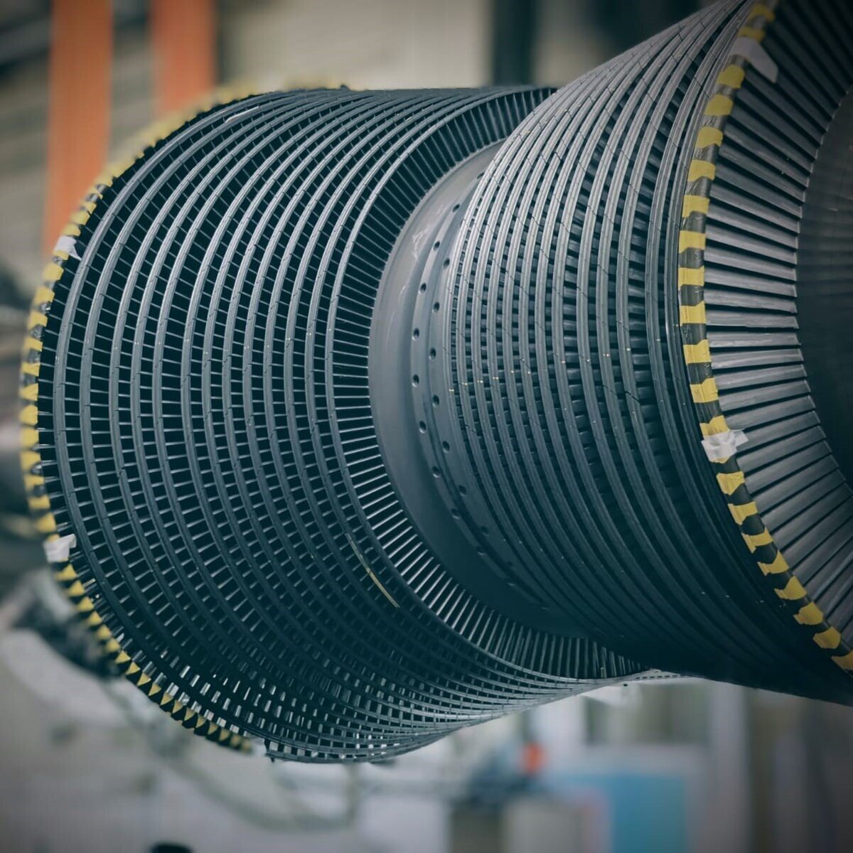 Close-up of a large industrial turbine rotor with numerous fine blades arranged in a spiral pattern. The rotor is seen in a factory environment, with the background blurred to focus attention on the detailed structure of the rotor.