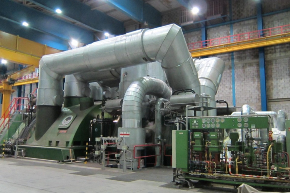 Large industrial machine with metal pipes and ducts, some parts painted green, in a factory setting with control panels, valves, and structural elements like beams and cranes.