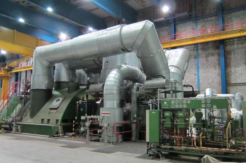 Large industrial machine with metal pipes and ducts, some parts painted green, in a factory setting with control panels, valves, and structural elements like beams and cranes.