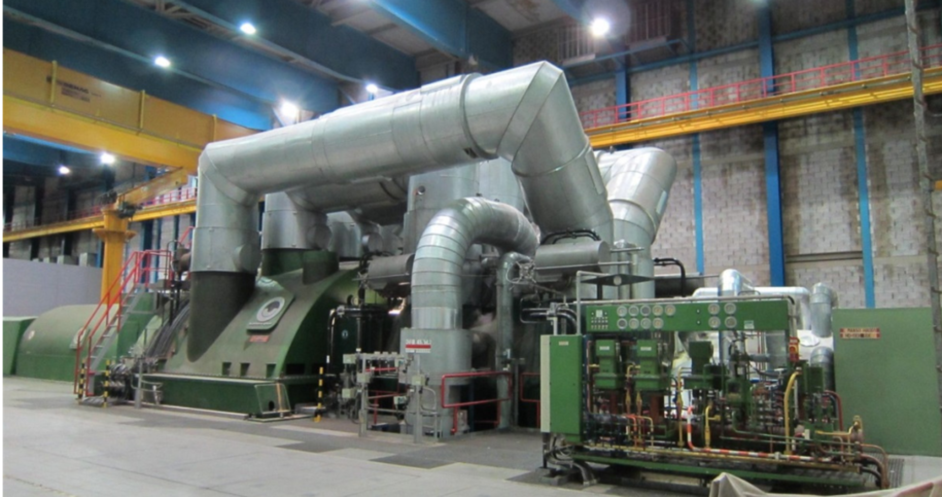Large industrial machine with metal pipes and ducts, some parts painted green, in a factory setting with control panels, valves, and structural elements like beams and cranes.