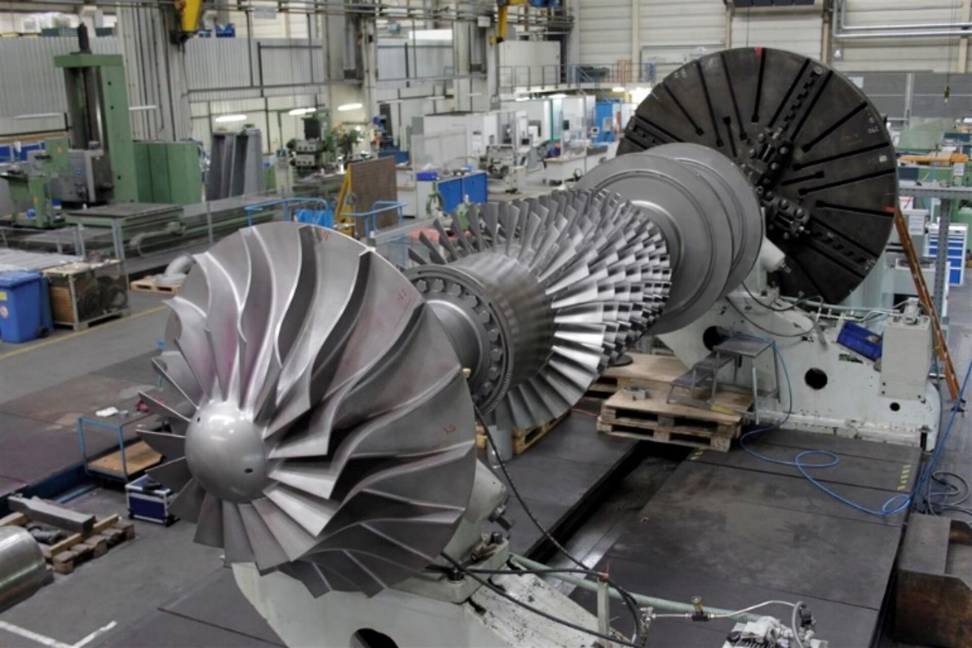 Large turbine rotor assembly in a factory hall, surrounded by machinery and equipment.