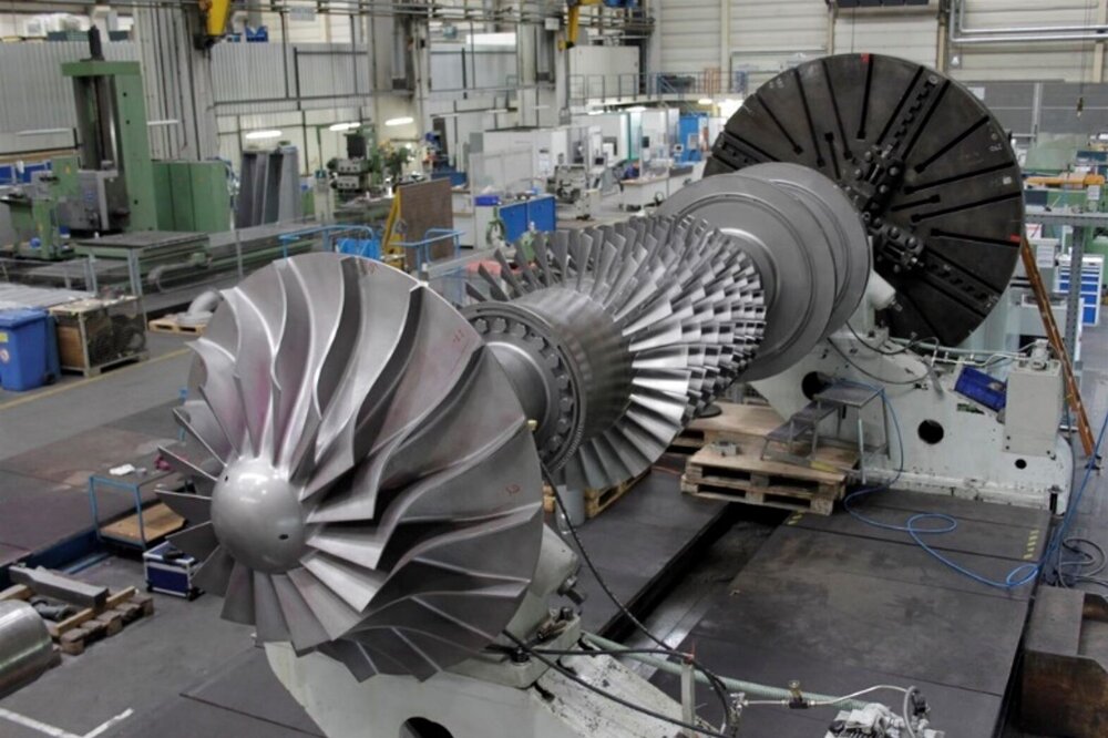 Large turbine rotor assembly in a factory hall, surrounded by machinery and equipment.